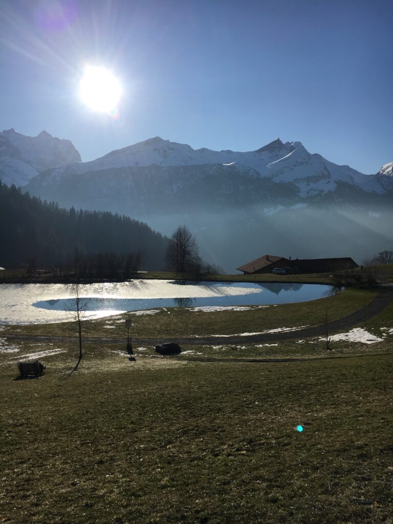 Winterlandschaft am Hasliberg mit kleinem Weiher, Sonnenspiegelung und Bergpanorama trotz wenig Schnee