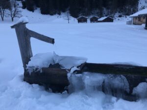 Vereister Brunnen auf der Schwarzwaldalp im Winter mit verschneiten Alphütten im Hintergrund