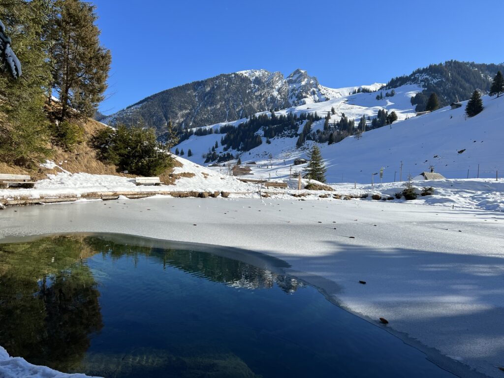 Blauseeli auf der Grimmialp im Winter, teilweise zugefroren, mit spiegelndem Wasser und verschneiten Bergen im Diemtigtal