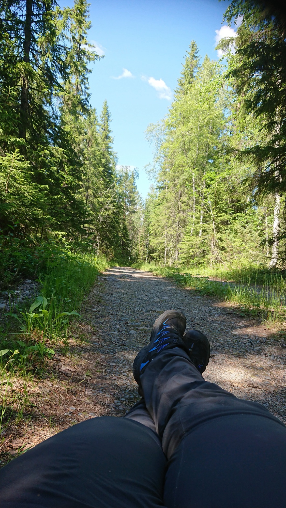 Pause beim Genusswandern auf einem ruhigen Waldweg – ausgestreckte Beine mit Wanderschuhen im Vordergrund, umgeben von grünem Wald und sommerlichem Licht.