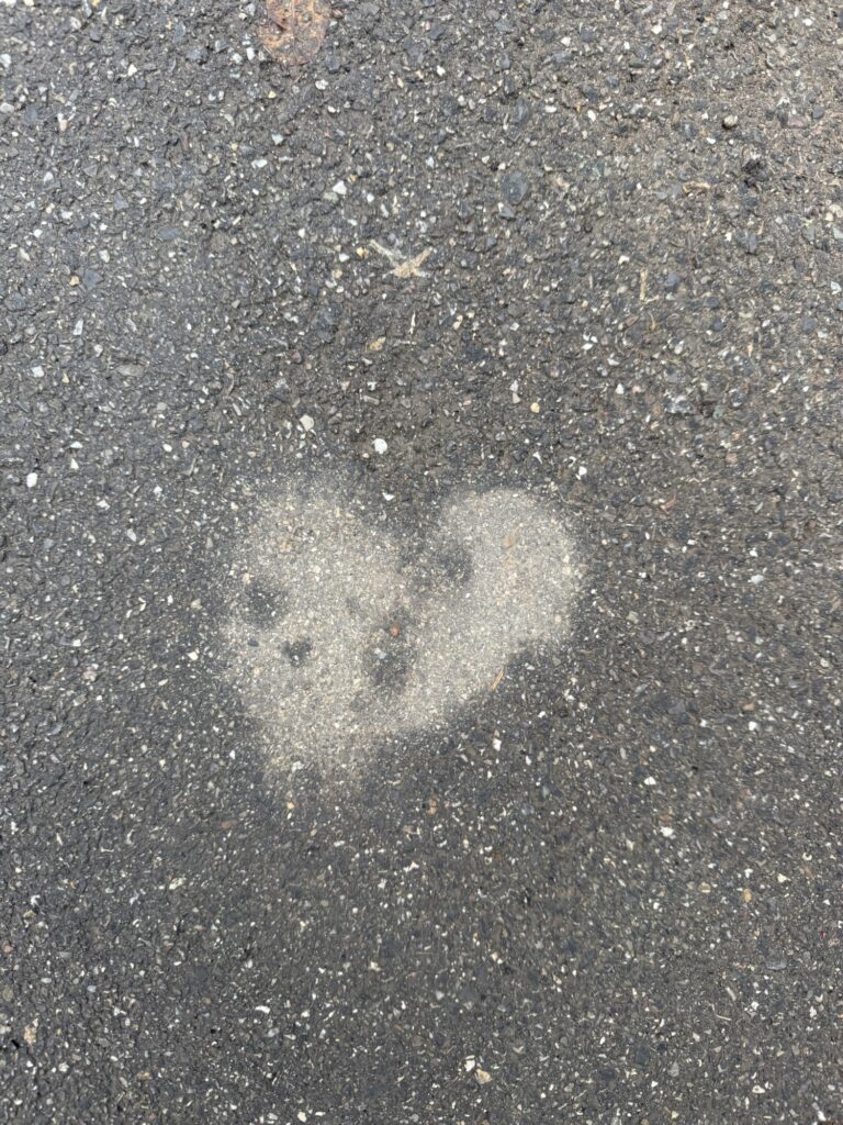 Ein herzförmiger Fleck aus trockenem Asphalt, umgeben von nasser Strasse – Liebe hebt sich selbst im Regen sichtbar hervor.