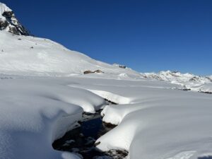 Winterlandschaft auf der Engstligenalp mit verschneiten Hügeln, kleinem Bach und Alphütten unter blauem Himmel