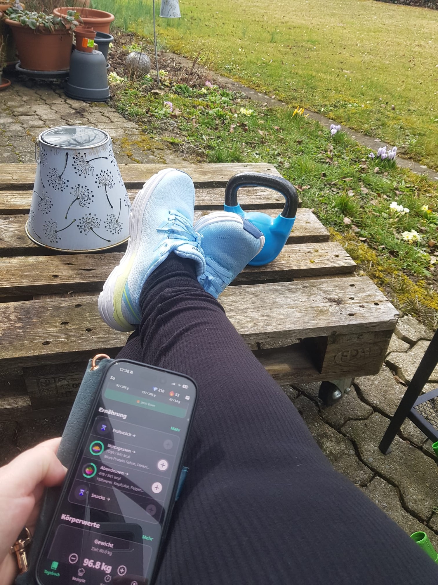 Tracking von Ernährung und Gewicht mit der Yazio-App nach dem Training im Garten – Sportschuhe, Smartphone und Kettlebell als Symbol für gesunde Routine und Fitness.