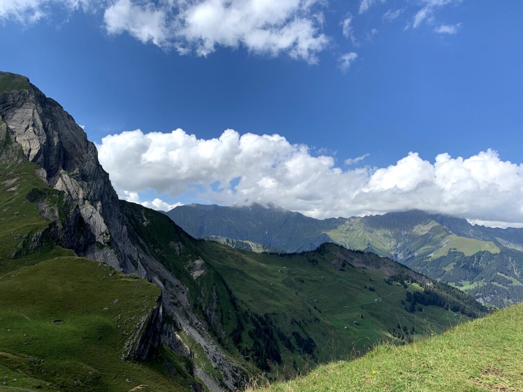 Panoramablick auf grüne Alpenlandschaft mit markanten Felswänden, Wanderwegen und sommerlichen Wolken über den Schweizer Bergen