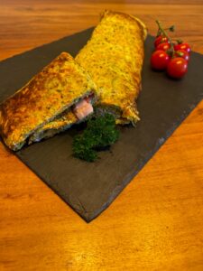 Goldbraun gebackene Gemüse-Ei-Rolle mit sichtbarer Füllung aus Schinken auf schwarzer Schieferplatte angerichtet. Dazu frische Petersilie und rote Cherrytomaten auf warmem Holztisch.